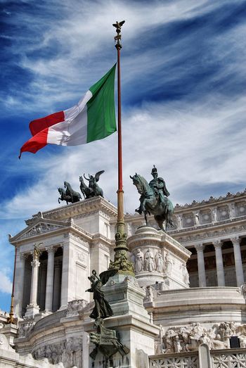 Low angle view of flag and statue at altare della patria against cloudy blue sky