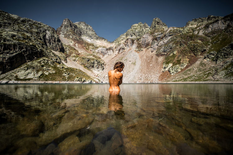 Back view of unrecognizable graceful female traveler standing in calm crystal transparent lake water against majestic rocky landscape in pyrenees mountains