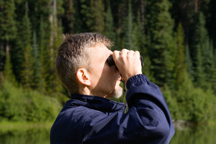 Side view of man looking through binoculars against trees