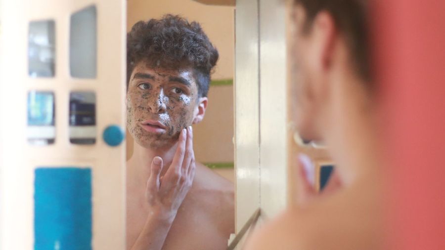 Shirtless man applying facial mask reflecting in mirror