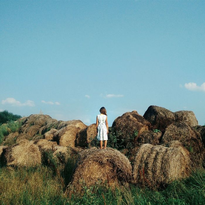 Rear view of young woman standing hay bale against blue sky