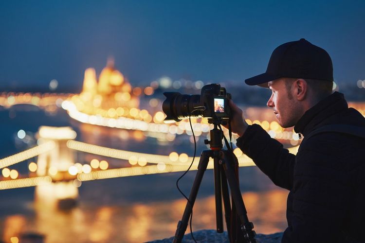 Man photographing illuminated chain bridge from camera in city at night