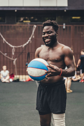 Happy shirtless man with sports ball standing at basketball court