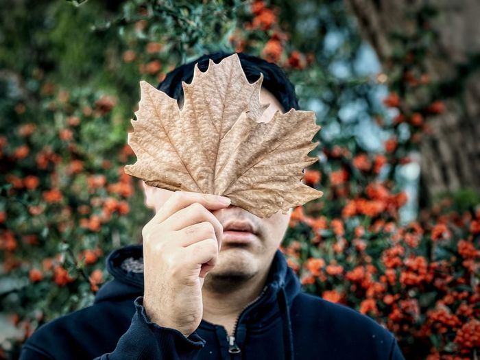 Midsection of man holding dried leaf against orange rowan berries and trees in autumn.