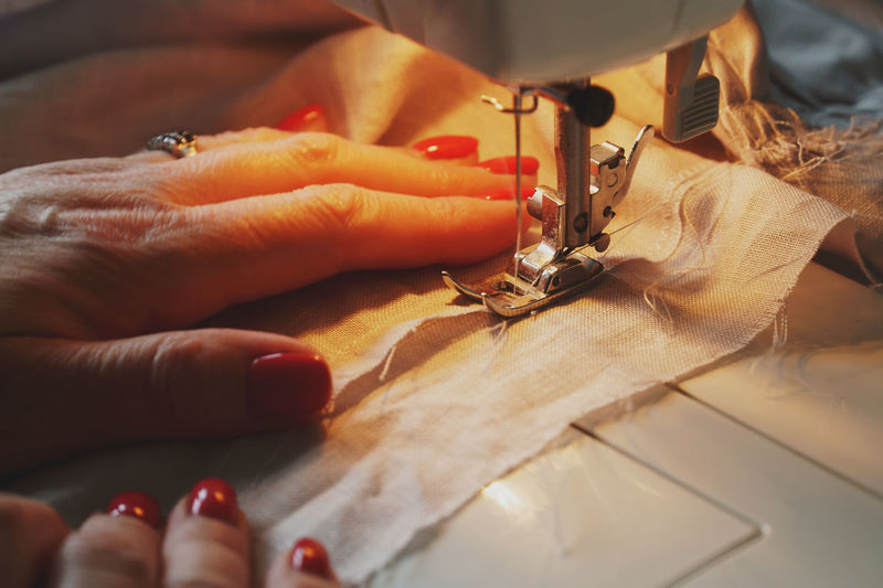 Photo of woman's hands with red nail polish in process of sewing linen dress using sewing machine 