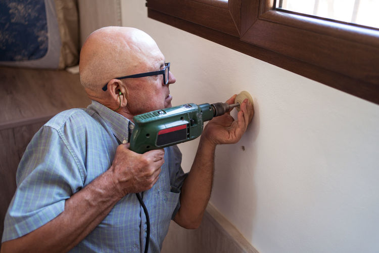 Concentrated old male in eyewear with electric screwdriver screwing plastic piece to wall in house