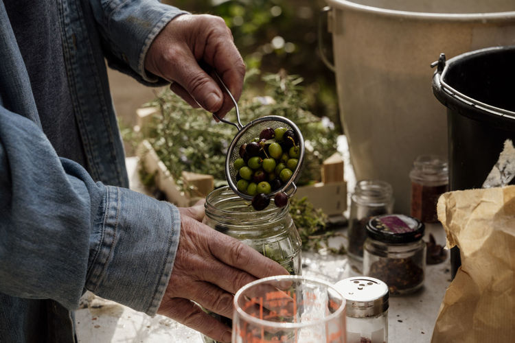 Senior man filling glass jar with olives from colander on table