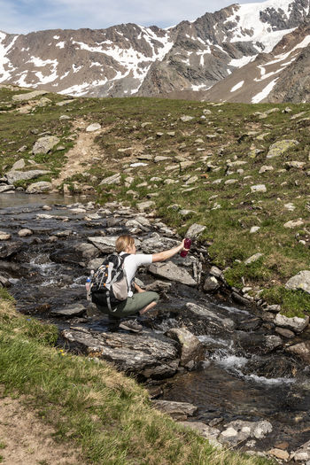 Rear view of woman crouching on rock by stream against mountains