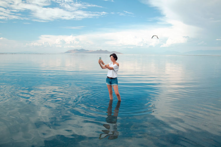 Woman in denim shorts and white t-shirt enjoying blue lake water. female using mobile phone .