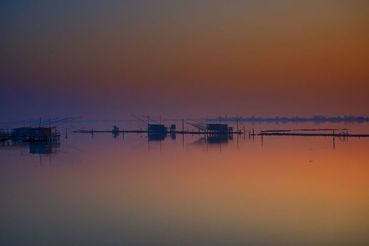 Fishing huts by stilts on sea against sky during sunset