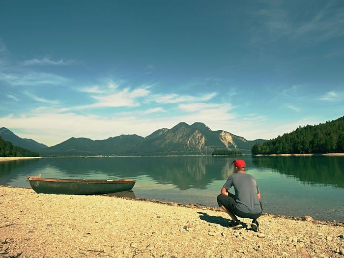 Man sitting on the stony bank of alpine mountain lake at anchored laminated boat.