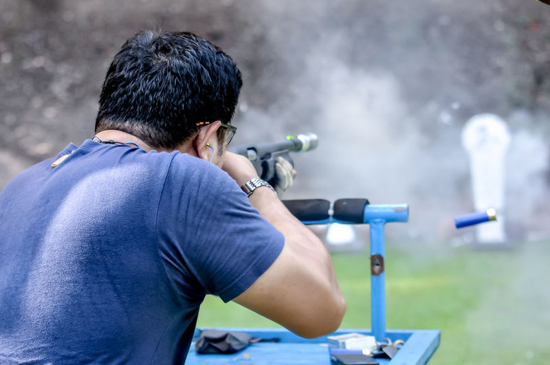 Rear view of man holding gun while aiming on target outdoors