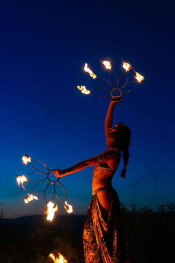 Side view of talented female fire dancer in costume standing with burning fire fans during performance on dark street in town