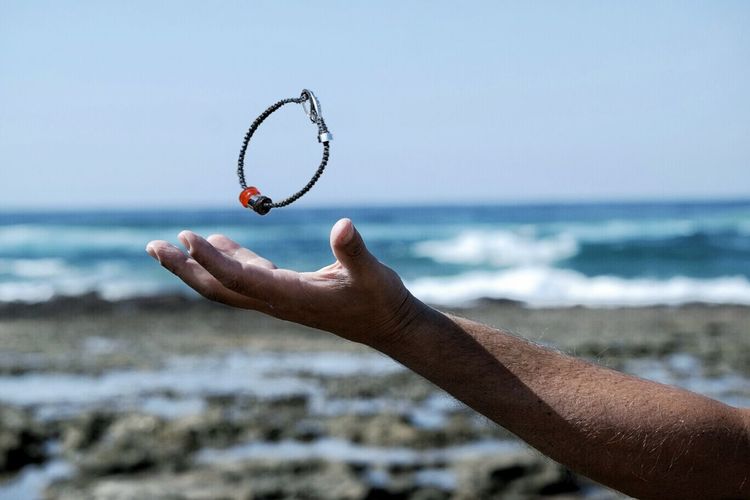 Cropped hand holding of man playing with bracelet against sky