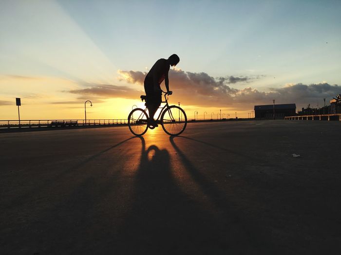 Silhouette man riding bicycle on street against sky during sunset