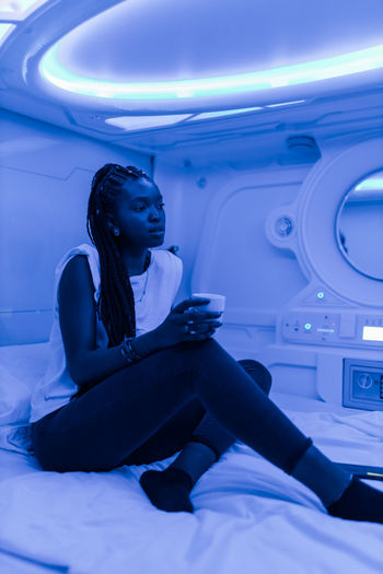 Tranquil black female sitting with cup of drink on bed in room of capsule hotel while resting during vacation