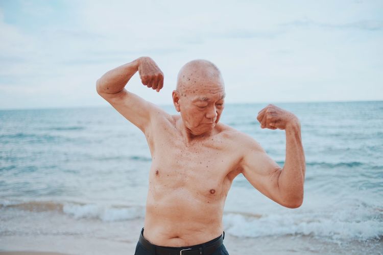 Shirtless senior man flexing muscles while standing at beach against sky during sunset