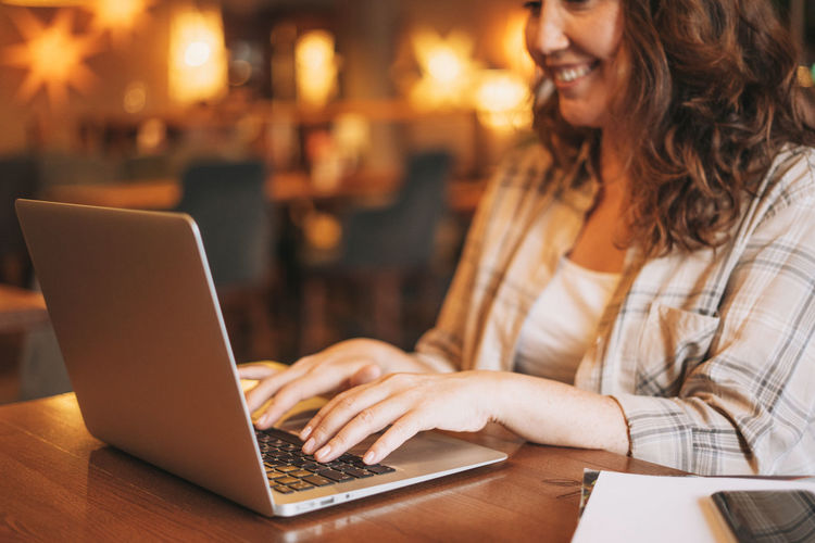 Crop photo of adult charming brunette woman in plaid shirt working with laptop at the cafe