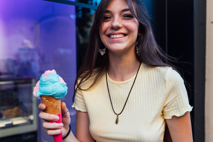 Crop cheerful young female in pendant and earrings with delicious gelato in waffle cone looking at camera on street