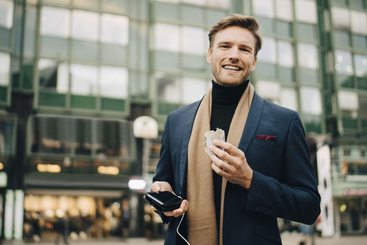 Portrait of smiling businessman with wrap sandwich standing in city
