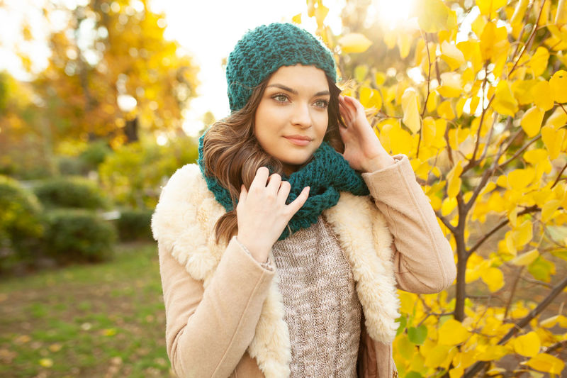 Beautiful young woman in warm clothing standing against autumn tree