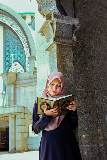 Young woman wearing hijab while reading holy book at mosque