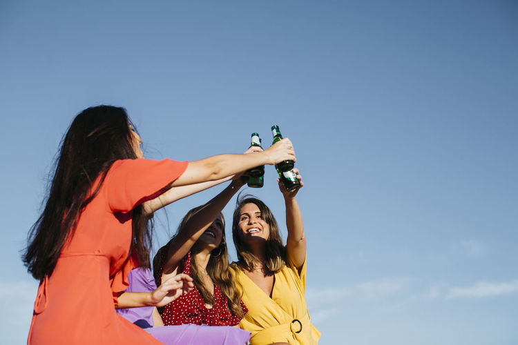 From below of group of joyful female friends toasting and clinking glass bottles of beer