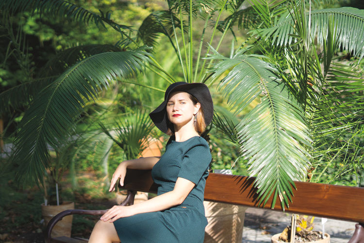 Portrait of woman wearing hat sitting on bench against tree