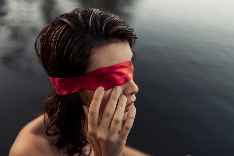 Side view of anonymous gentle female tourist in red blindfold touching face against water in daylight