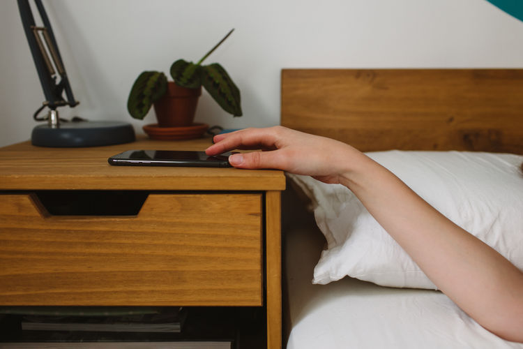 Cropped unrecognizable person hands turning off alarm on mobile phone while lying in bed after awakening in cozy bedroom
