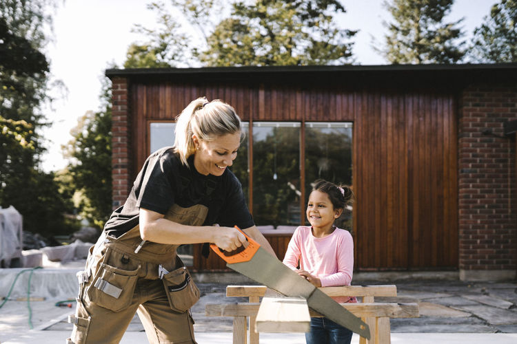 Smiling daughter looking at mother cutting plank with hand saw against house