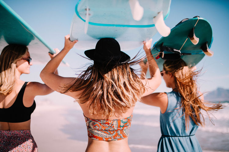 Rear view of female friends carrying surfboards on head at beach