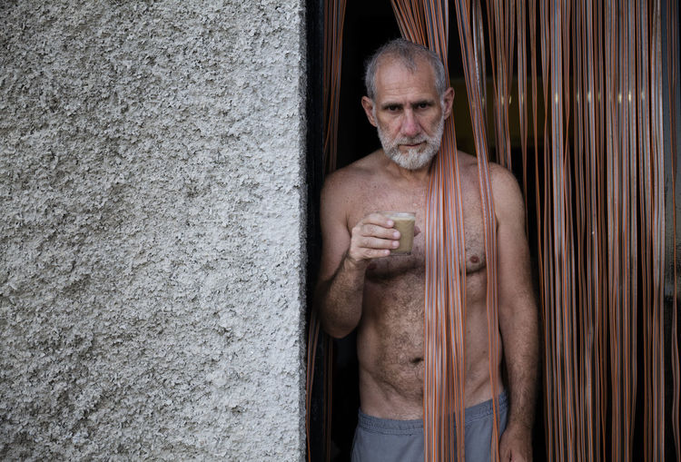 Portrait of shirtless man having coffee on door standing against wall