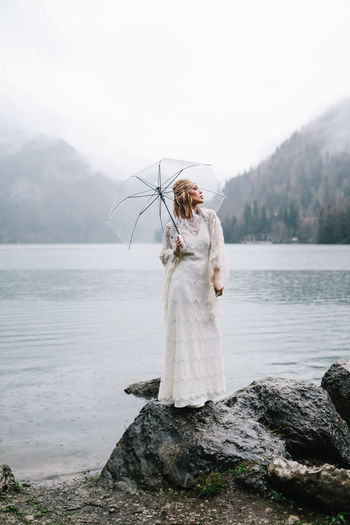 A young woman in a white lace wedding dress stands in the rain among the sea and mountains