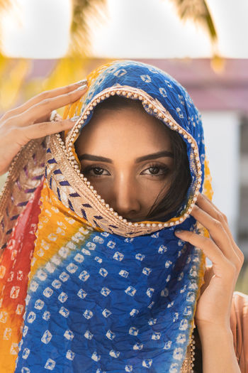 Young ethnic indian female in traditional colorful headscarf looking at camera while standing against blurred tropical yard