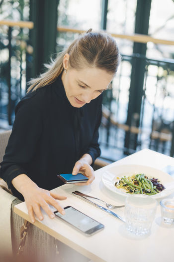 Confident businesswoman using smart phone while sitting with credit card at cafeteria in office