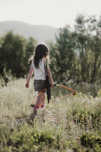 Rear view of girl walking while holding bow and arrow on grassy field