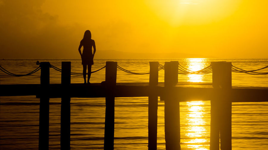 Silhouette woman standing on pier over sea against orange sky