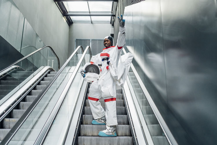 Cheerful astronaut gesturing peace sign while climbing on escalator