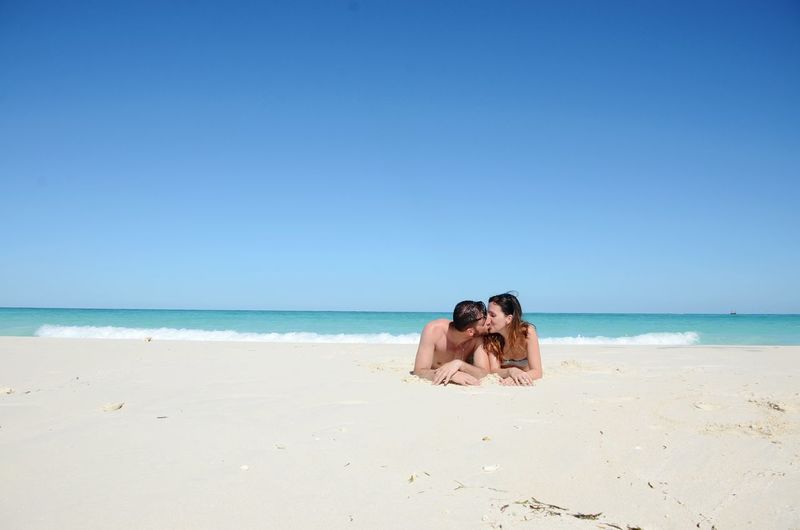 Couple kissing while lying on sand at beach against sky during sunny day