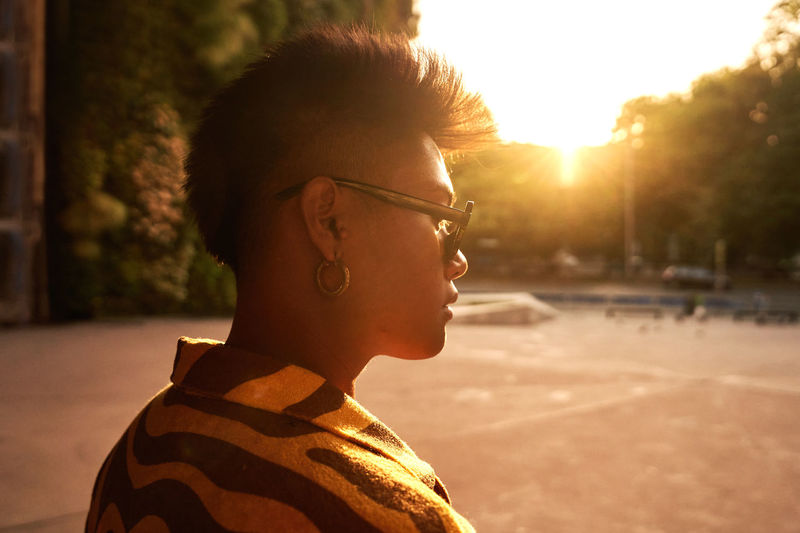 Side view of cool asian male in trendy shirt and earrings wearing sunglasses while standing in park in back lit of sunset light
