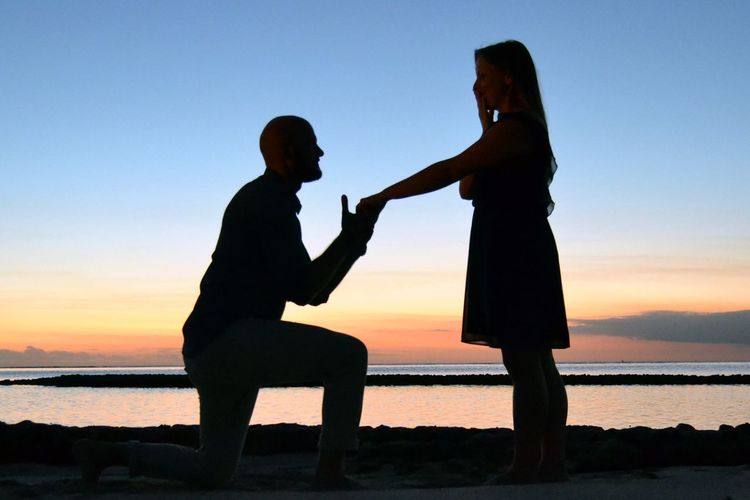 Silhouette man proposing woman while holding hand at beach against sky during sunset