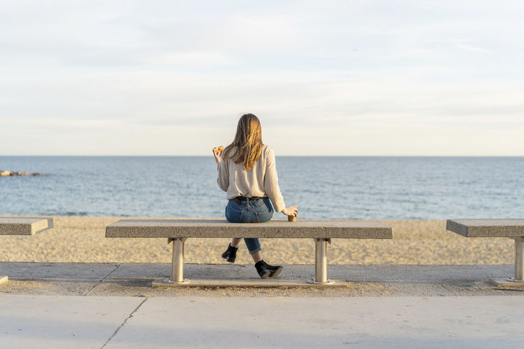 Young woman looking at sea while sitting on bench enjoying sunset