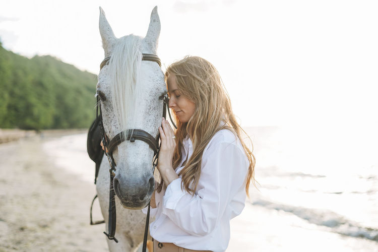 Young long hair woman in white shirt riding white horse on seascape background