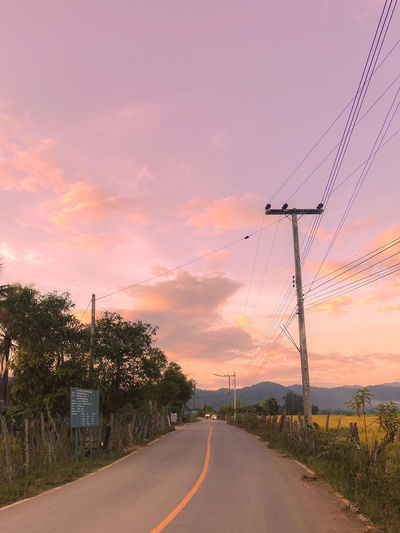 Road by electricity pylon against sky during sunset