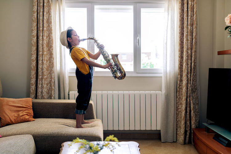 Side view of barefoot child with closed eyes playing saxophone while standing on couch at home in daytime