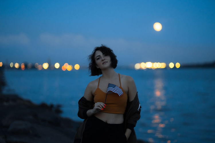 Portrait of woman holding flag while standing at beach against sky at dusk