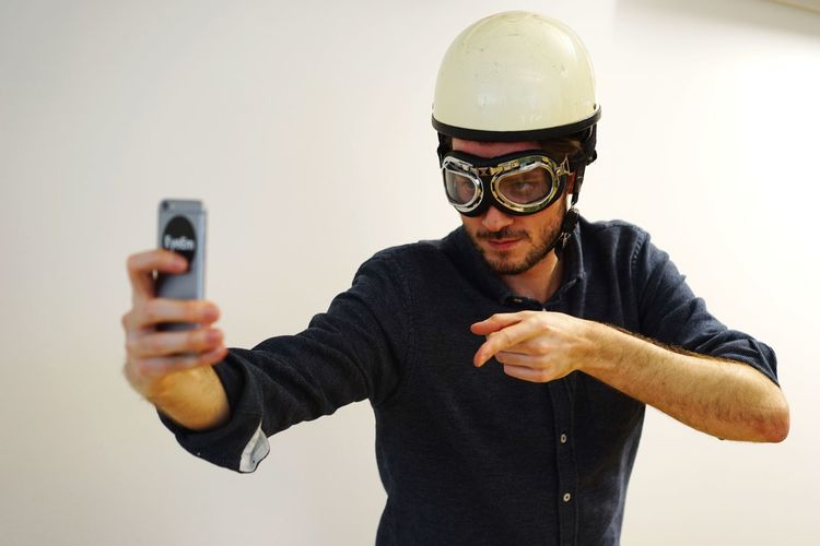 Young man in aviator glasses and helmet taking selfie against white wall