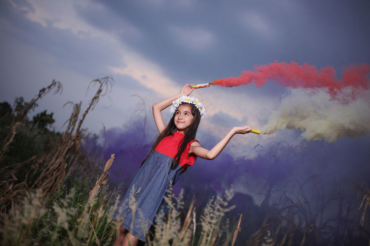 Smiling cute girl holding distress flares while standing against sky at dusk