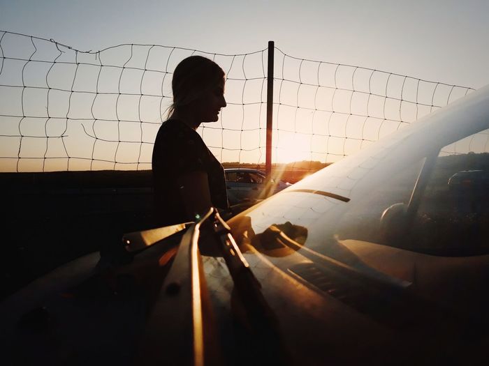 Side view of silhouette woman standing by car against sky during sunset